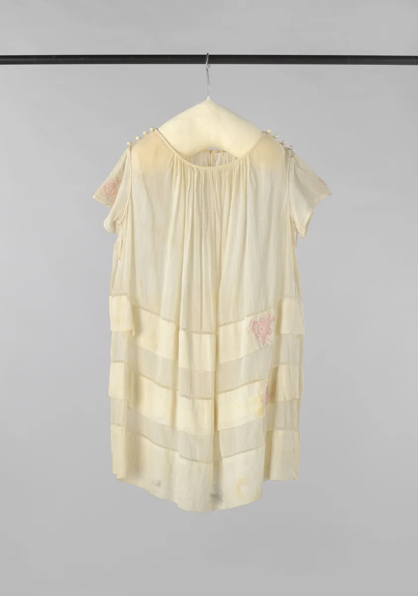 Kinderjurk met korte mouw, plooien vanuit de hals en vier gehaakte bollen op de schouder van wit katoen, versierd net ajourranden en twee stroken met roze geborduurde bloemen by anonymous, other, 1900-1925