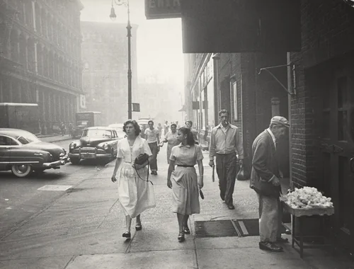 It is 8:30 and the street is beginning to come to life. The fruit peddler who keeps his fruits in the garage has arranged his tray. On the left Connie Damiani is on her way to her job at Plymouth Toy Co. by Robert Frank, photograph, 1951