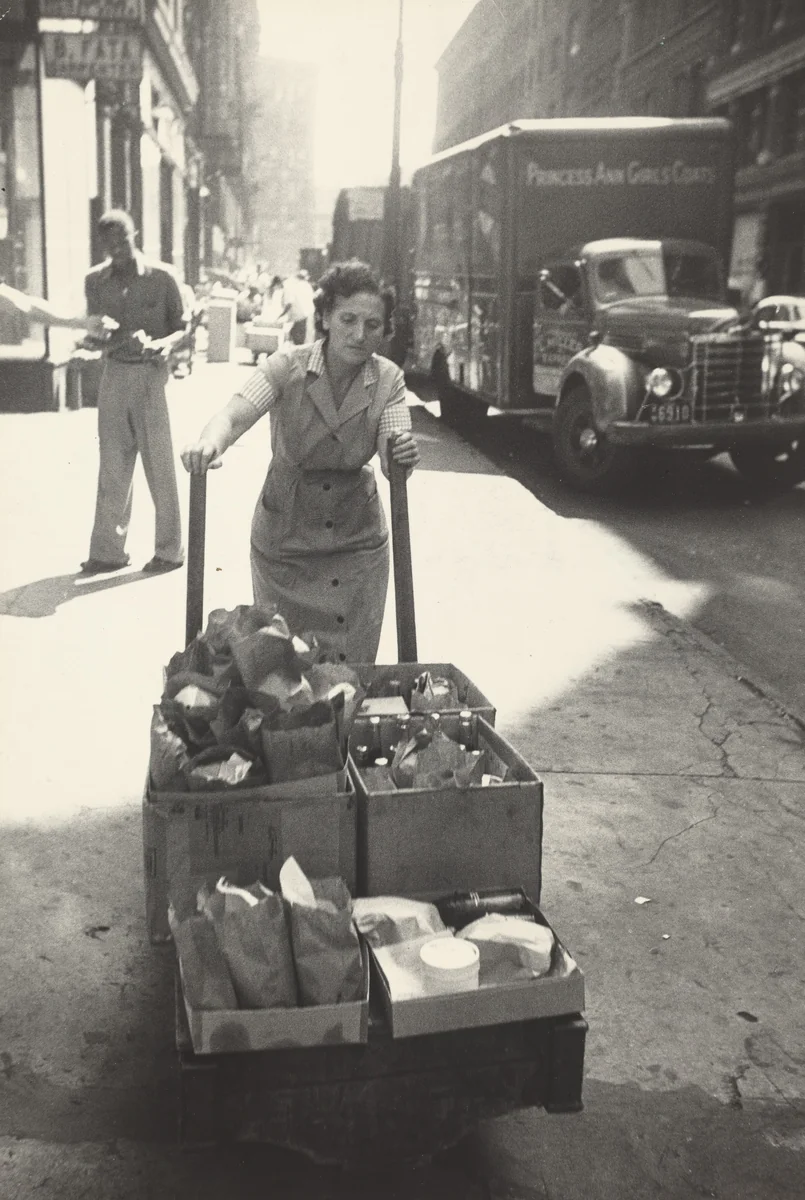 Just before noon Lena Kansler delivers orders to the building at the west corner of the block. Her husband Benny runs the luncheonette. by Robert Frank, photograph, 1951