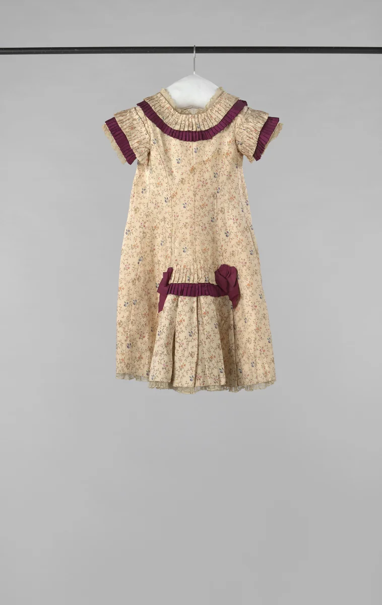 Een feestjurkje van bedrukte creme zijde met bloemtakjes, opgemaakt met karmijnrode strikjes en stroken. Het schootje is van glasbatist, en onder de rokzoom komt een Valenciennes-kant te voorschijn by anonymous, other, 1880-1885