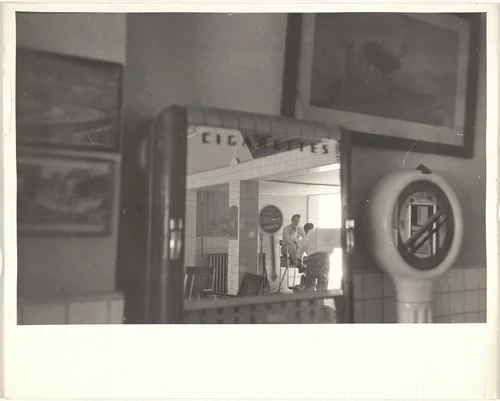 Shining shoes, reflection in cigarette machine mirror--New Mexico by Robert Frank, photograph, 1955