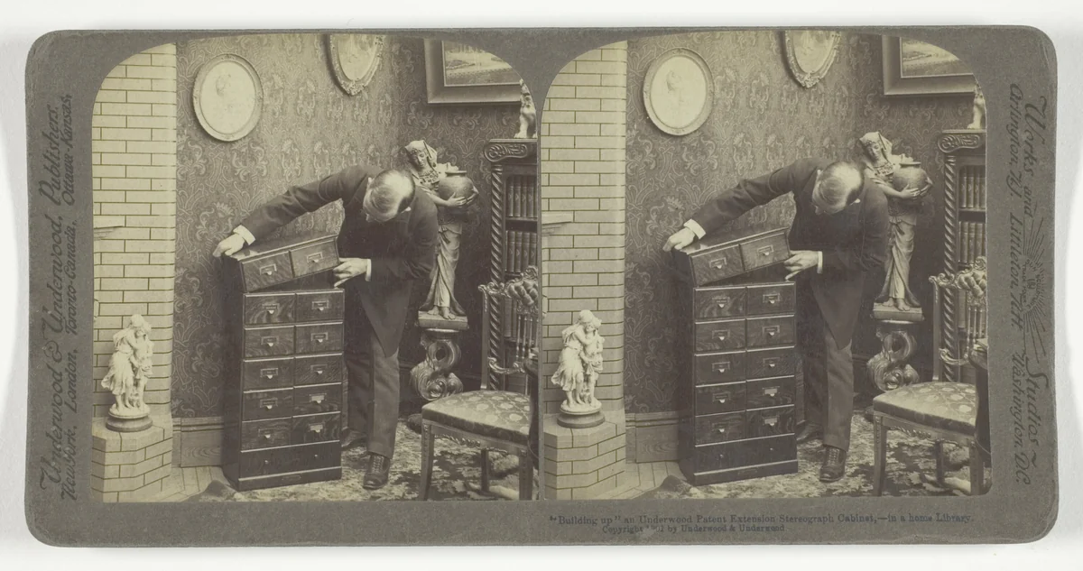 "Building up" an Underwood Patent Extension Stereograph Cabinet, - in a home Library. by Underwood & Underwood, photograph, 1901