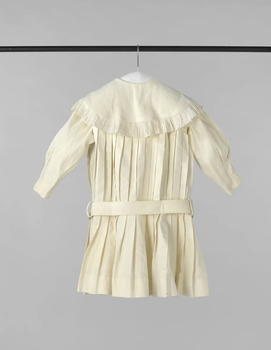 Een kleuterjurk van in plooien genaaide piqué, voorzien van ronde liggende kraag met batisten ruches en een stoffen ceintuur by anonymous, other, 1875-1880